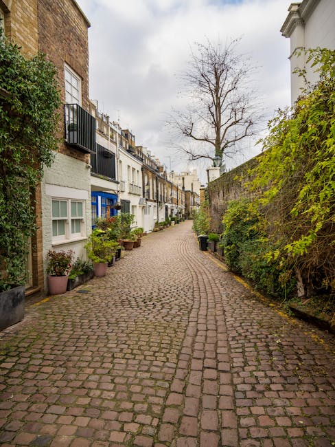 A residential street lined with parked cars on both sides, with the foreground showing dark asphalt pavement. In the middle ground, several low-branched trees with pink blossoms extend over the pavement, creating a canopy of light pink flowers contrasting with the dark branches. The row of Victorian-style terraced houses with white facades, decorative details, and sash windows line the street on either side, partially obscured by the trees. The sky above is partly cloudy with patches of blue visible, and the scene is illuminated by natural daylight, suggesting a springtime setting. The overall scene depicts a quiet urban environment where easy access for private waste collection or rubbish clearance services like those offered by [COMPANY_NAME] may be visually associated with the curb-side configuration and residential nature of the area.