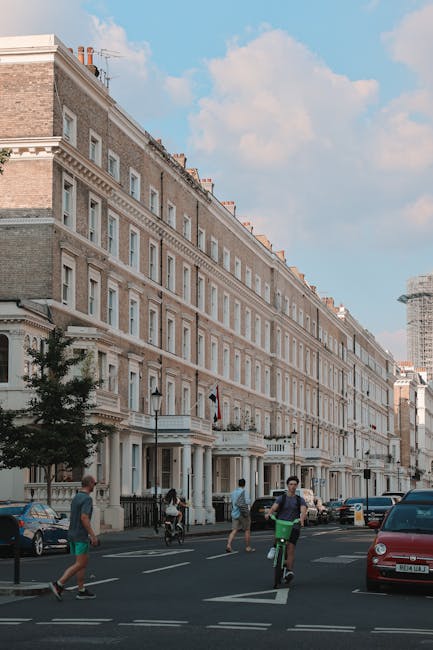 The image depicts a bustling street scene in South Kensington, featuring a row of ornate, multi-storey residential buildings with classic architecture, including decorative cornices and large sash windows, constructed from beige and light brown bricks with white detailing. The buildings are positioned along a paved road with parked cars lining the curb and a few moving vehicles visible in the background. In the foreground, several pedestrians are walking on the sidewalk, while a man on a green bicycle rides past. A small tree is planted along the pavement, and the sky above is partly cloudy with patches of blue, creating a bright, natural lighting environment. The scene reflects an urban area where private waste management services, such as those offered by rubbish clearance providers like [COMPANY_NAME], may facilitate alternative rubbish removal solutions, exemplified by the presence of waste collection or clearance activities typical in such residential settings.
