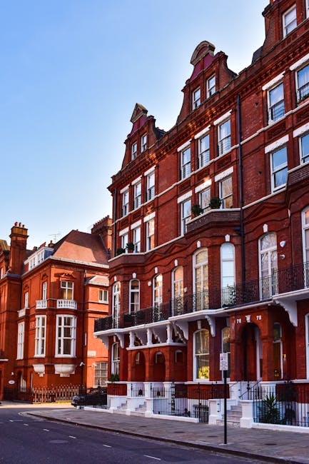 The image depicts a bustling street scene in South Kensington, featuring a row of ornate, multi-storey residential buildings with classic architecture, including decorative cornices and large sash windows, constructed from beige and light brown bricks with white detailing. The buildings are positioned along a paved road with parked cars lining the curb and a few moving vehicles visible in the background. In the foreground, several pedestrians are walking on the sidewalk, while a man on a green bicycle rides past. A small tree is planted along the pavement, and the sky above is partly cloudy with patches of blue, creating a bright, natural lighting environment. The scene reflects an urban area where private waste management services, such as those offered by rubbish clearance providers like [COMPANY_NAME], may facilitate alternative rubbish removal solutions, exemplified by the presence of waste collection or clearance activities typical in such residential settings.