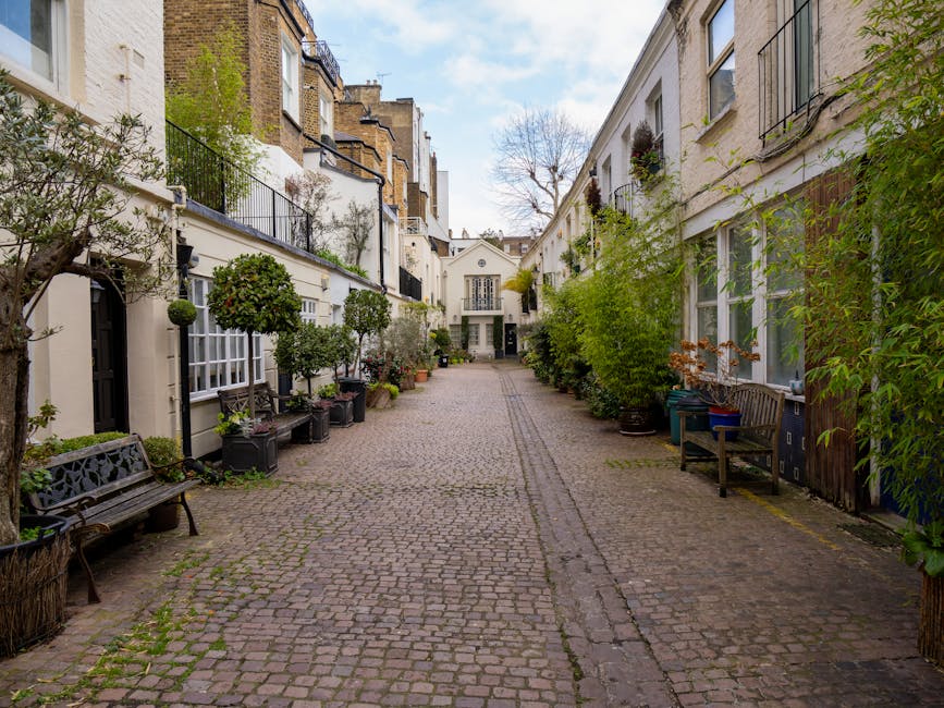 A narrow residential cobblestone alleyway bordered by two- and three-storey white-painted buildings on either side. The buildings feature large sash windows with black railings and flower boxes, some with potted plants and small trees placed along the ground level and on window sills. On the left side, a black metal bench is situated beneath a potted shrub, while on the right, a wooden bench and a blue recycling bin are positioned near a door. The alley is lined with various potted plants, including small trees, bushes, and flowering pots, creating a lush, green appearance. The cobblestone surface shows slight unevenness with narrow, darker stone pathways running down the middle. The sky overhead is partly cloudy, allowing soft natural light to illuminate the scene, emphasizing the textured surfaces of the buildings and greenery. The setting appears tranquil and well-maintained, indicative of a typical private courtyard or alley that might be involved in a local rubbish removal service or private waste handling context, such as that provided by Rubbish Clearance South Kensington.