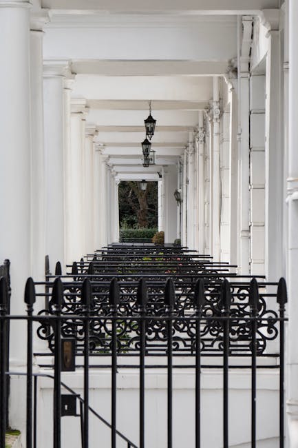 A long, covered pathway with a white-painted wooden ceiling supported by evenly spaced beams and columns. The corridor is enclosed by a black wrought iron fence at the front, featuring decorative patterns and a small gate, with a doorbell or intercom mounted beside it. Inside, three hanging lantern-style light fixtures are attached to the ceiling, aligned along the length of the corridor. The flooring appears to be a light-colored, smooth surface, possibly stone or concrete. At the far end, a garden with a large tree and some shrubbery is visible, providing a natural backdrop to the otherwise structured, architectural setting. The scene is illuminated by natural light, creating a bright atmosphere suitable for pedestrian passage, possibly part of a private estate or residential property. This type of setting is typical for on-site clearance or private rubbish collection, with the clean and well-maintained environment indicating ongoing or recent maintenance work by a waste management service.
