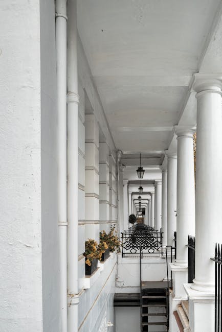A narrow residential cobblestone alleyway bordered by two- and three-storey white-painted buildings on either side. The buildings feature large sash windows with black railings and flower boxes, some with potted plants and small trees placed along the ground level and on window sills. On the left side, a black metal bench is situated beneath a potted shrub, while on the right, a wooden bench and a blue recycling bin are positioned near a door. The alley is lined with various potted plants, including small trees, bushes, and flowering pots, creating a lush, green appearance. The cobblestone surface shows slight unevenness with narrow, darker stone pathways running down the middle. The sky overhead is partly cloudy, allowing soft natural light to illuminate the scene, emphasizing the textured surfaces of the buildings and greenery. The setting appears tranquil and well-maintained, indicative of a typical private courtyard or alley that might be involved in a local rubbish removal service or private waste handling context, such as that provided by Rubbish Clearance South Kensington.