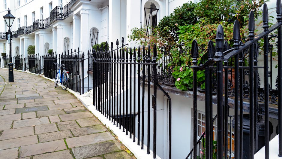 A row of white terraced townhouses with black wrought iron railings and decorative fencing along a paved sidewalk. The buildings have black window frames and balcony railings, with some covered in climbing green plants and pink flowers. In the foreground, a blue bicycle is leaning against one section of the fence. The paved walkway consists of large rectangular stone slabs, some showing signs of weathering, leading past the front entrances. An underground staircase is visible behind the iron fence, indicating an entrance below ground level. The scene is outdoors during daylight, with natural light illuminating the residential area, suggesting the setting relates to private or independent waste management options for property upkeep or rubbish clearance, as might be handled by services like those at rubbishclearancesouthkensington.co.uk.