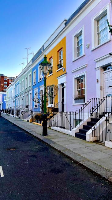 A row of colorful terraced houses in South Kensington, featuring facades painted in pastel shades of blue, yellow, and lavender, with white window frames and decorative trims. The houses are two stories high, with small front gardens enclosed by black wrought-iron railings and steps leading up to their entrances. A classic black street lamp is positioned in the foreground, mounted on a short stone base, with a glass lantern and a decorative finial on top. The pavement consists of concrete slabs, with a narrow asphalt road running alongside the houses, which appears clean and well-maintained. The sky is clear, suggesting a bright and sunny day. The scene reflects an urban residential environment that might be associated with private waste collection or alternative rubbish removal services such as on-site clearance, often used as an alternative to local authority disposal, aligning with the context of rubbish removal services.
