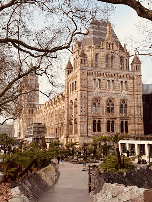 The image depicts a historic, multi-story building constructed from light brown and beige stone with intricate masonry details, arched windows, and pointed turrets, set against a clear sky with leafless tree branches extending across the top. In the foreground, there is a pedestrian pathway bordered by natural stone rock formations and landscaped green plants, including ferns and small trees, leading towards the building. Several people are visible walking along the pathway, which is lined with metal bollards. To the right, modern glass and steel structures are partially visible, contrasting with the historic architecture. The overall scene suggests an urban environment with a mix of old and new architecture, surrounded by landscaping that may be part of a public or private outdoor space. This setting is consistent with urban areas where private or independent waste collection services, such as rubbish clearance, are employed to manage waste disposal in conspicuous, high-traffic areas. The image’s composition, lighting, and clarity align with a professional setting aimed at highlighting architectural and environmental details associated with rubbish removal or site clearance activities.