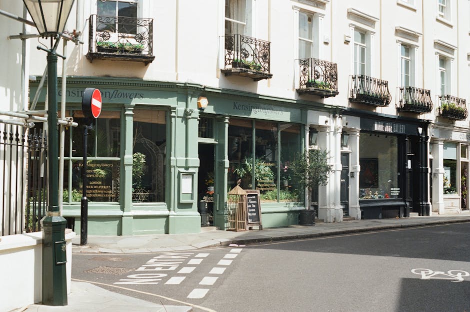 The image depicts a row of three-connected commercial buildings on a city street, with the ground floors occupied by retail shops and the upper floors featuring residential-style windows and small balconies with black wrought-iron railings and potted plants. The lowest level includes large front windows displaying greenery and products, framed by pastel green and darker black facades, providing a contrast in textures and finishes. In front of the shops, there is a sidewalk with a black street lamp on the left, a bollard, and a no-entry traffic sign mounted on a pole near the corner. Visible markings in the paved road include a bicycle lane symbol and direction arrows. The lighting appears natural, with soft daylight casting minimal shadows, contributing to a calm and orderly urban scene. This setting is consistent with a central London area, where private commercial premises are characteristic, aligning with services such as independent rubbish clearance providers like rubbishclearancesouthkensington.co.uk operating in the vicinity to manage waste from retail and residential properties.